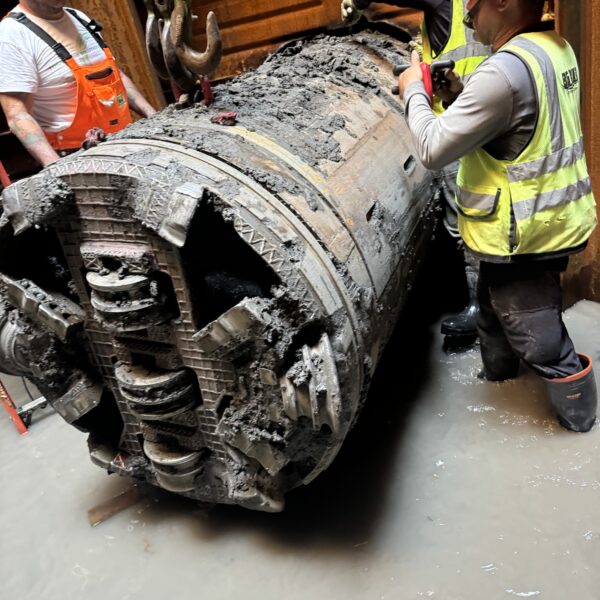 Three construction workers wearing hard hats and high-visibility vests inspect a large, burnt, mud-covered cylindrical industrial component in a wet work area.
