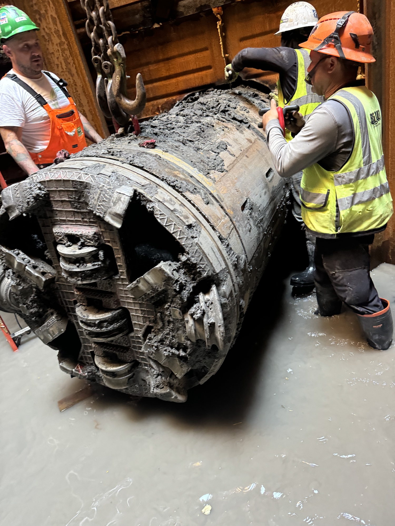 Three construction workers wearing hard hats and high-visibility vests inspect a large, burnt, mud-covered cylindrical industrial component in a wet work area.