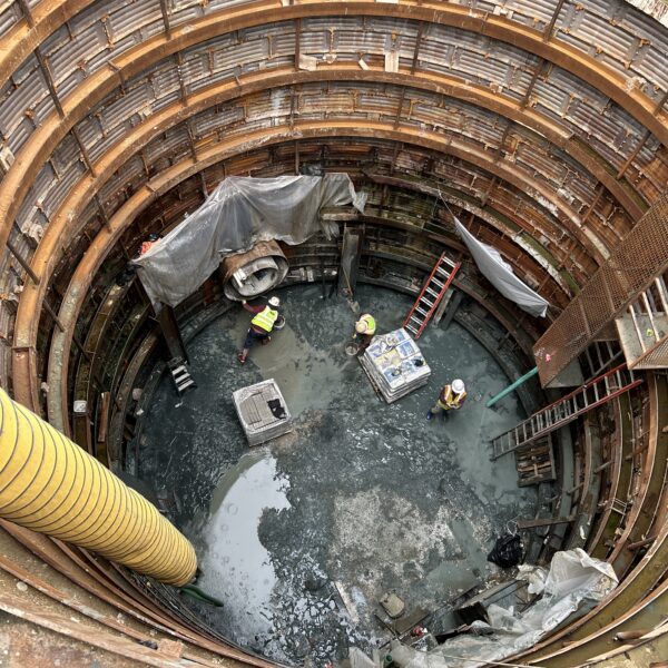 2369A233-61A1-4FC9-94B4-6ED075E88206 - Big River Trenchless & GeoServices View down into a rusted circular excavation shaft with stacked steel formwork rings; workers in high-visibility clothing at the bottom near muddy water.