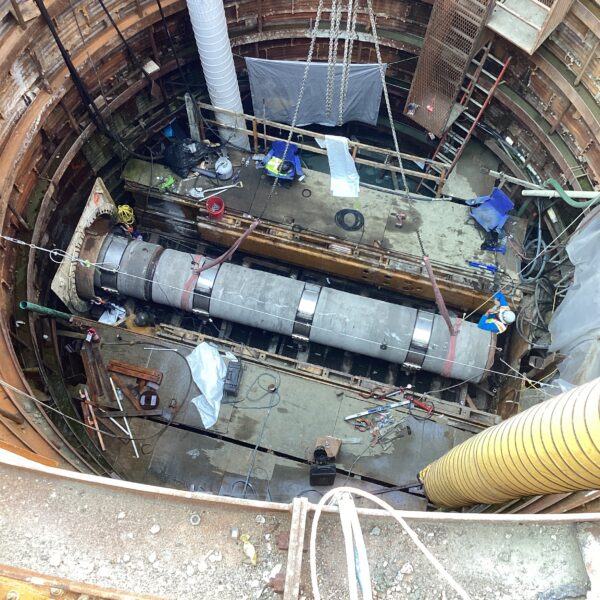 2ADDB24C-026D-4613-B046-A8D87470C78A - Big River Trenchless & GeoServices Circular excavation shaft under construction with concentric steel rings and scaffolding, chained hoist above, and cluttered work floor with hoses and tools.