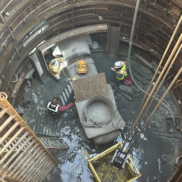 2BB3B31A-DA2E-4562-9507-DD3660AD563A - Big River Trenchless & GeoServices Top-down view of workers in a deep circular shaft, surrounded by steel rings and scaffolding, with concrete segments and water at the bottom.