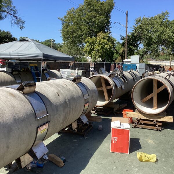 4DD73E56-CBAF-457F-BC57-820E25B1BFA0 - Big River Trenchless & GeoServices Industrial yard with large concrete pipes on wooden supports under a gray canopy, blue sky above.