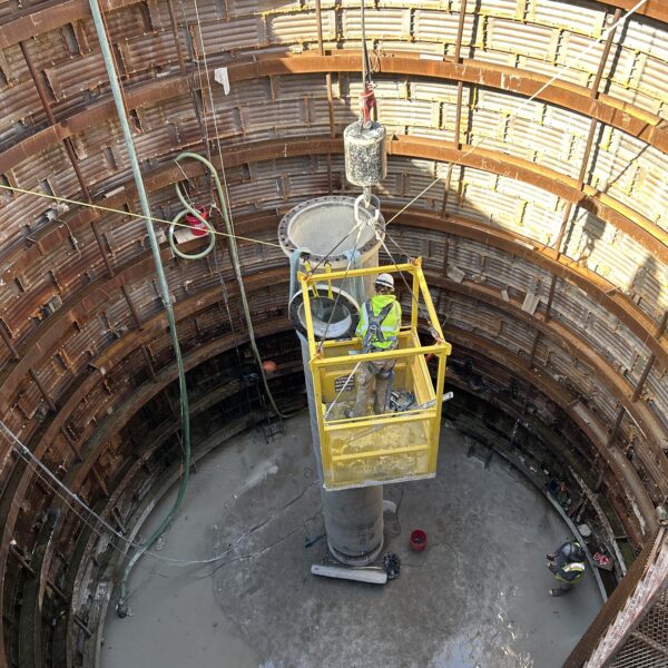 516C269C-DE6C-4CA5-91BD-705594B758A6 - Big River Trenchless & GeoServices Worker in a yellow suspended platform inside a circular steel-sheeted trench, lowering a heavy bucket into muddy water.