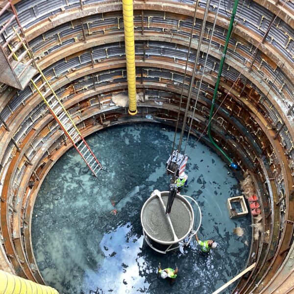 Procore - Big River Trenchless & GeoServices A deep circular excavation shaft with stacked metal rings, yellow hoses, and workers in high-visibility gear at the bottom over a muddy, water-filled base.