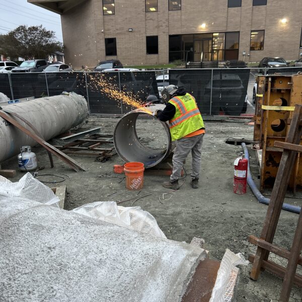 97EA6231-0BDD-427B-A8C6-BA124A50549C - Big River Trenchless & GeoServices Construction worker in a high-visibility vest and helmet grinding metal, sparks flying, at a fenced-out site with large concrete pipe pieces nearby.