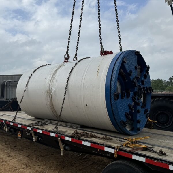 E602C9D6-E820-4199-B79A-C555C846AD96 - Big River Trenchless & GeoServices Large white cylindrical industrial component with blue ends on a flatbed trailer, suspended by crane chains during loading.