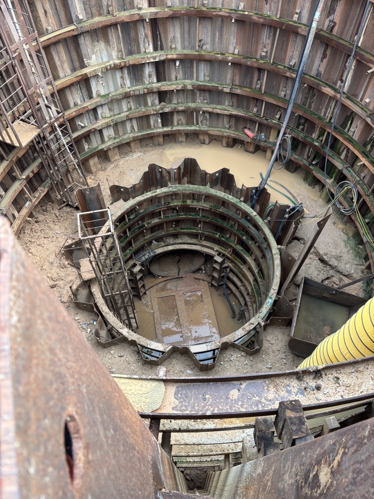 Deep circular excavation with multiple concentric steel sheet piles, ladders, and muddy water at the bottom of a construction shaft
