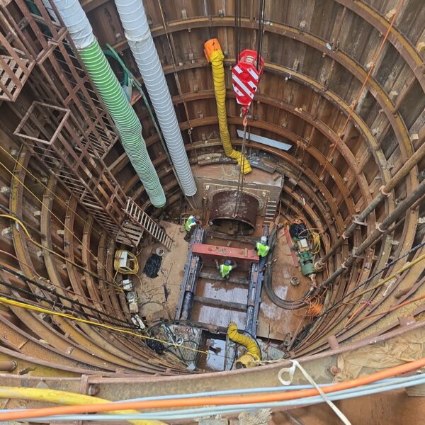 IMG_2026_07_06_17_12_02_819 - Big River Trenchless & GeoServices Deep circular underground shaft under construction, with timber/steel ring walls, large yellow and gray ducting, and workers in hi‑vis gear on a platform below.