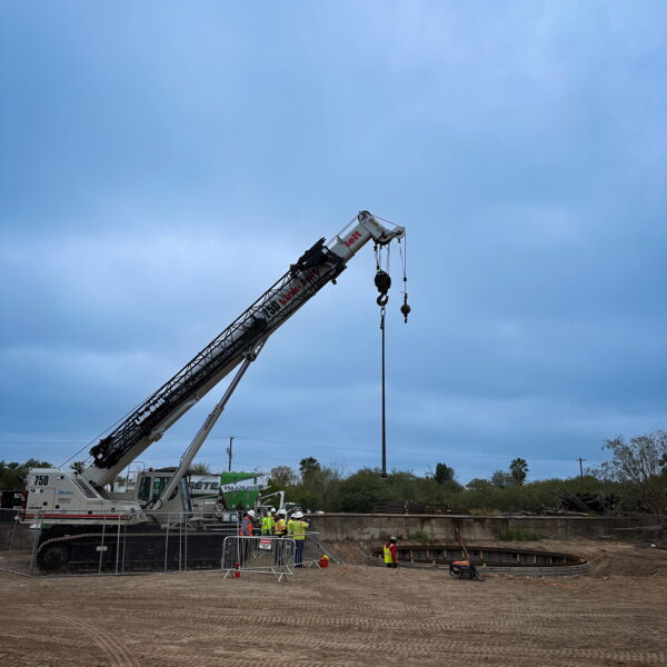 ZacateCreekWWTP-Img1 - Big River Trenchless & GeoServices Crane lifts a long arm over a construction site with workers in safety vests near a circular excavation pit surrounded by fencing and dirt ground.