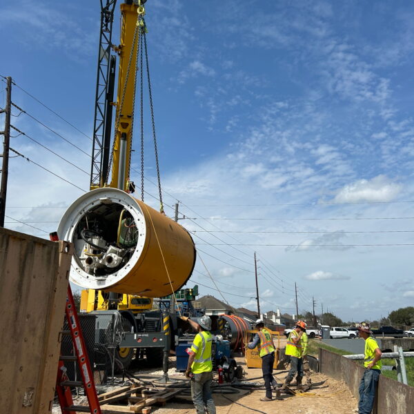 ZacateCreekWWTP-Img11 - Big River Trenchless & GeoServices Construction crew at a site with a large yellow drilling rig lowering a cylindrical component into a trench area, safety gear on workers visible