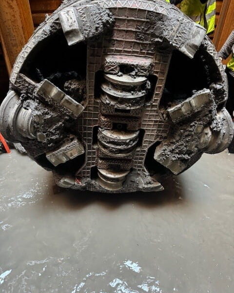 ZacateCreekWWTP-Img6 - Big River Trenchless & GeoServices Large muddy tunnel-boring machine cutter head with worn teeth, being moved by workers in safety gear.