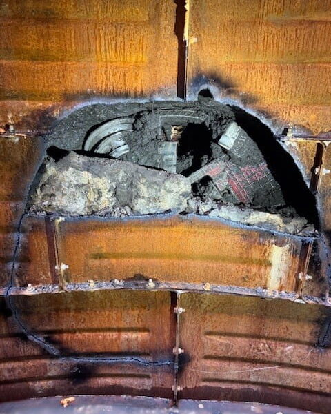 ZacateCreekWWTP-Img8 - Big River Trenchless & GeoServices Charred wooden ceiling with a large circular hole exposing dusty debris and damaged electrical conduit; soot and melted insulation around the opening.