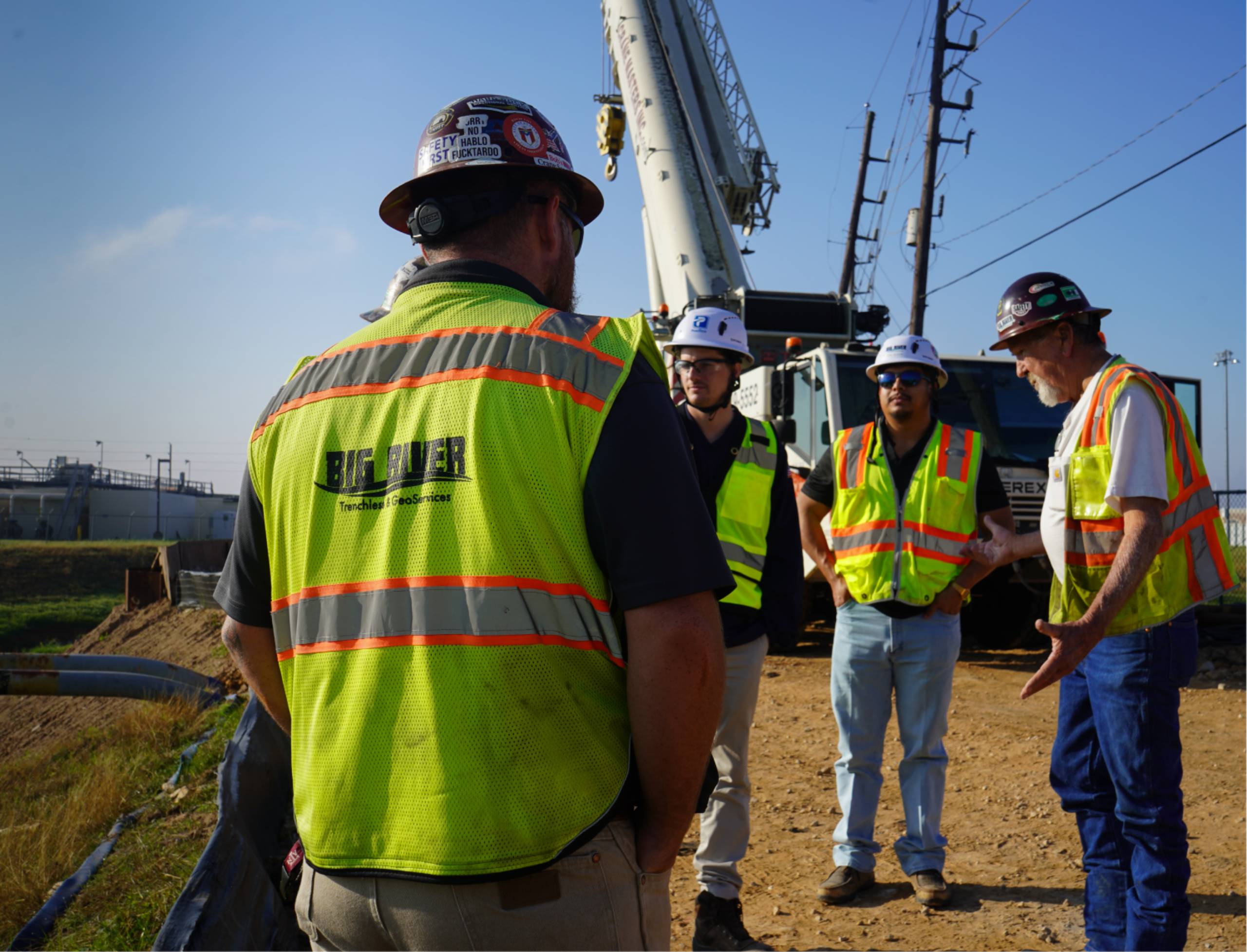 Group of construction workers in neon safety vests and hard hats standing on a dirt site with a large crane in the background, discussing a project.