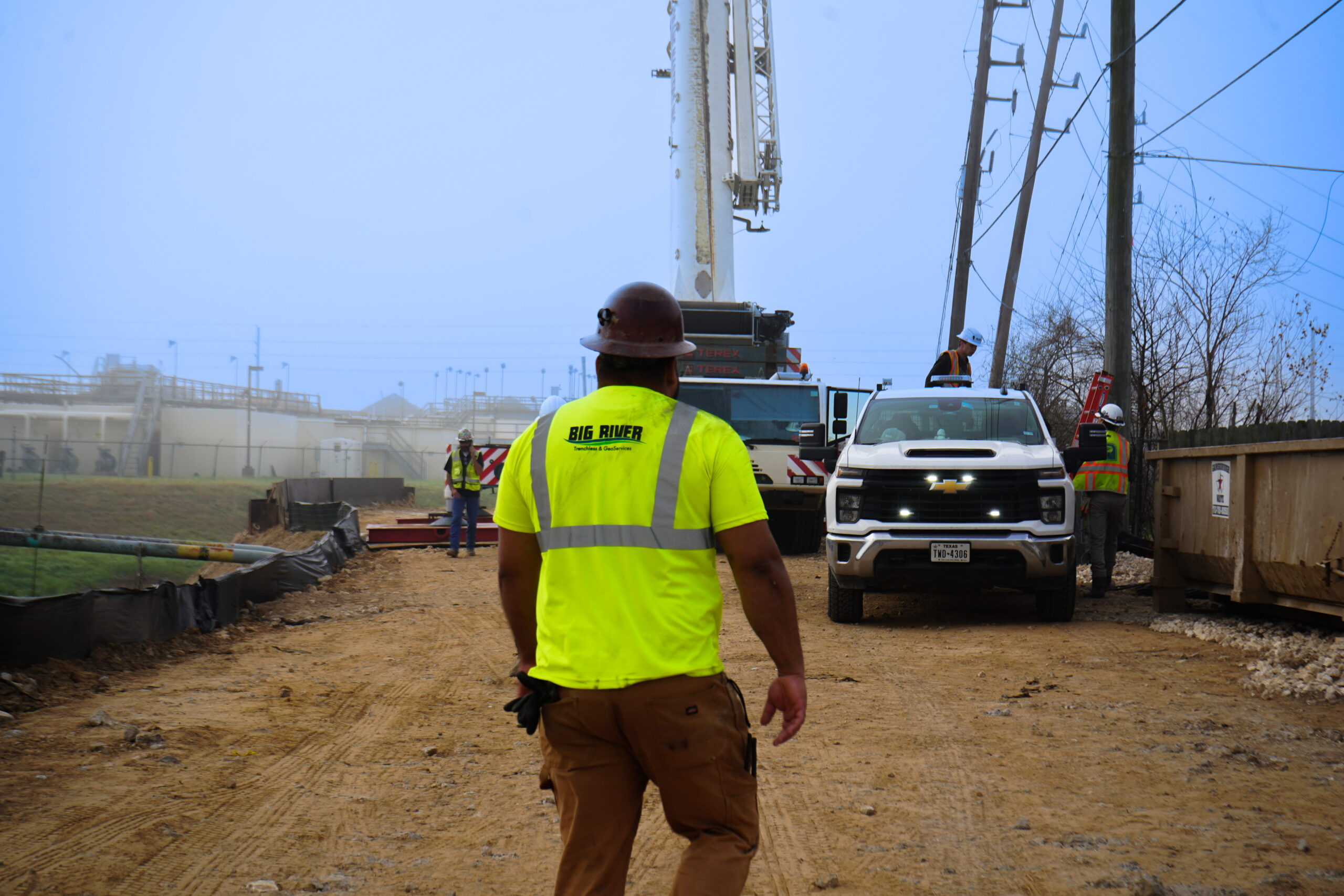 Construction crew at a worksite with a white truck, power lines, and a tall equipment tower in the background; worker in a neon yellow vest in foreground.