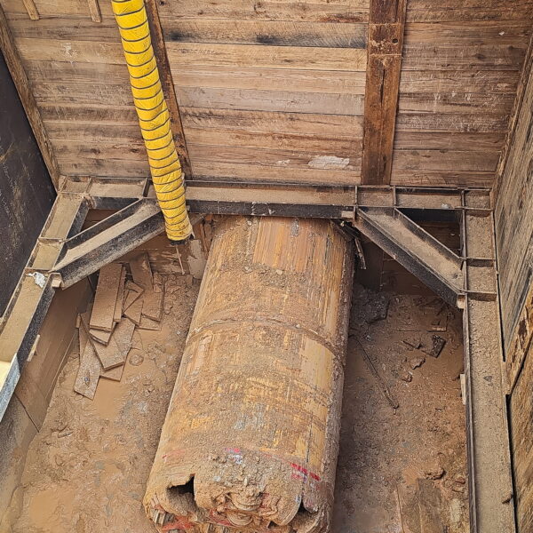 WHCRWA-Contract-64-Img10 - Big River Trenchless & GeoServices Construction trench with a large dusty pipe and a yellow spiral-wrapped conduit, surrounded by wooden supports and scattered debris on the muddy floor