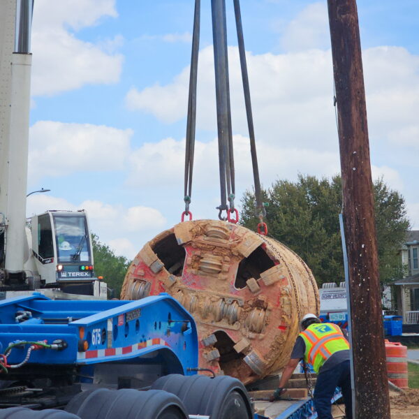 WHCRWA-Contract-64-Img12 - Big River Trenchless & GeoServices Massive cylindrical tunnel segment being lowered onto a blue trailer by cables, at a construction site with a crane and worker nearby in safety gear.