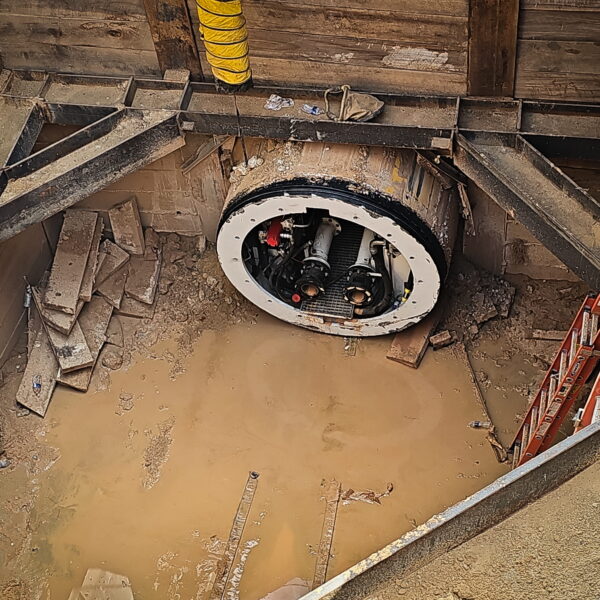 WHCRWA-Contract-64-Img14 - Big River Trenchless & GeoServices Construction excavation with a large circular pipe入口 in a muddy pit, surrounded by wooden formwork, a yellow vent hose, and orange ladders far right.