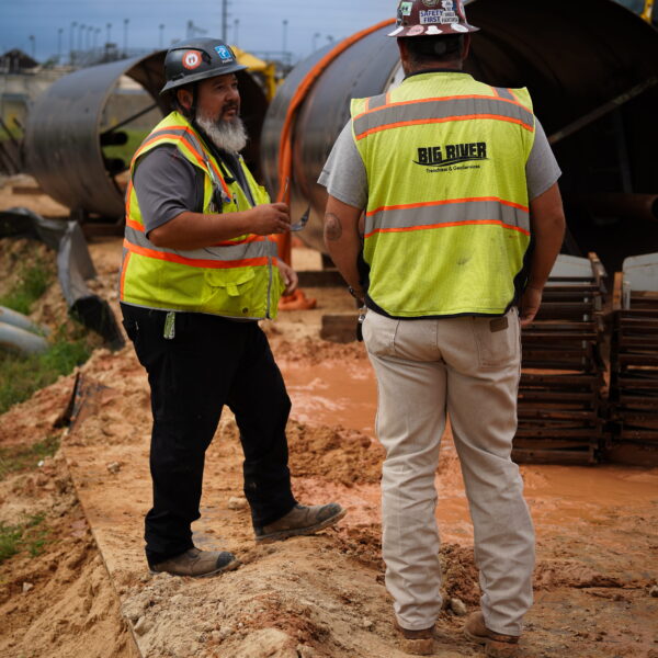 WHCRWA-Contract-64-Img15 - Big River Trenchless & GeoServices Two construction workers wearing high-visibility vests and hard hats stand near a large curved metal pipe on a muddy construction site.
