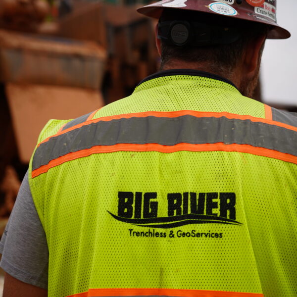 WHCRWA-Contract-64-Img17 - Big River Trenchless & GeoServices Back view of a construction worker in a neon yellow safety vest with orange reflective stripes; vest reads 'BIG RIVER' on the back, helmet covered in stickers; blurred construction site in the background.