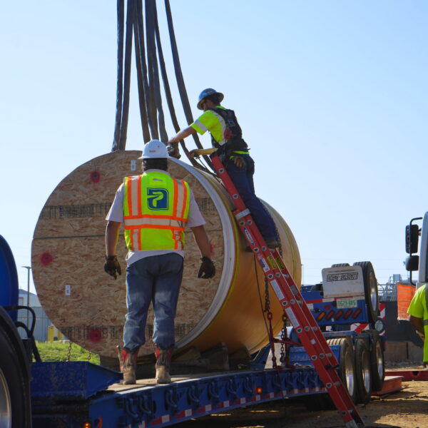 WHCRWA-Contract-64-Img5 - Big River Trenchless & GeoServices Two construction workers in safety vests and hard hats stand on a trailer beside a large wooden spool, with crane cables coming down.