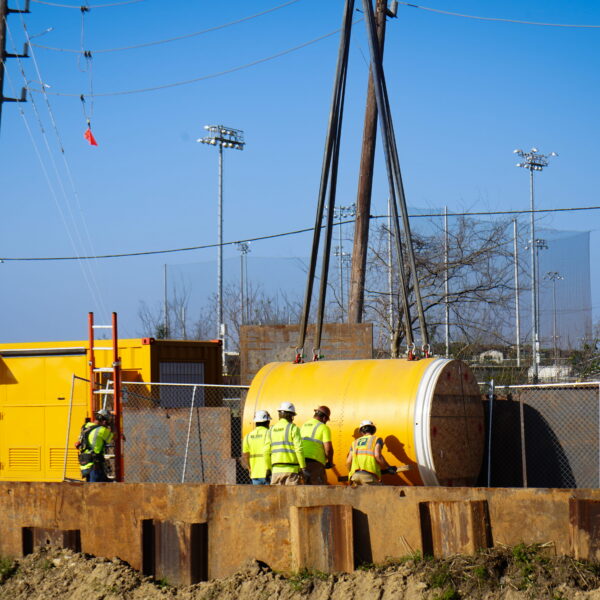 WHCRWA-Contract-64-Img6 - Big River Trenchless & GeoServices Construction workers in yellow safety vests and hard hats stand near large yellow industrial equipment on a fenced construction site with rusted steel beams and power lines against a clear blue sky.