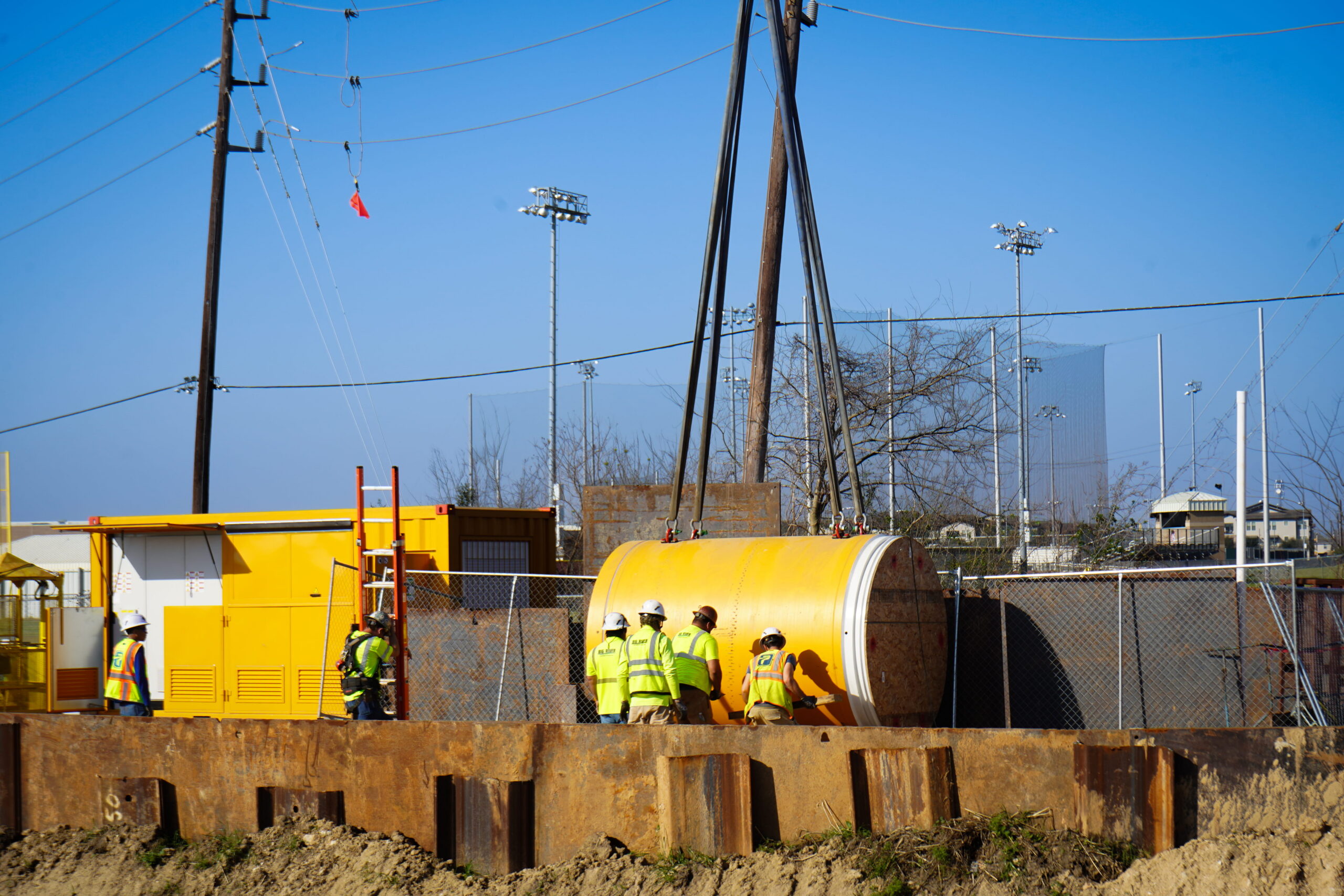 Construction workers in yellow safety vests and hard hats stand near large yellow industrial equipment on a fenced construction site with rusted steel beams and power lines against a clear blue sky.