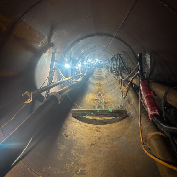 WHCRWA-Contract-64-Img7 - Big River Trenchless & GeoServices Interior of a cylindrical tunnel under construction with exposed pipes, cables, and temporary lighting along the walls